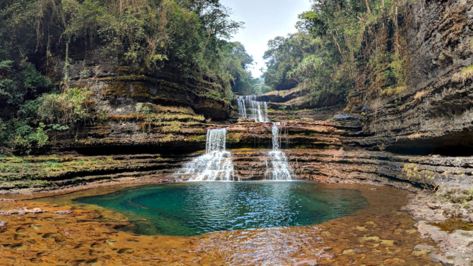 A three-tiered waterfall dropping over layered brown rock into a round blue-green pool in a forested gorge