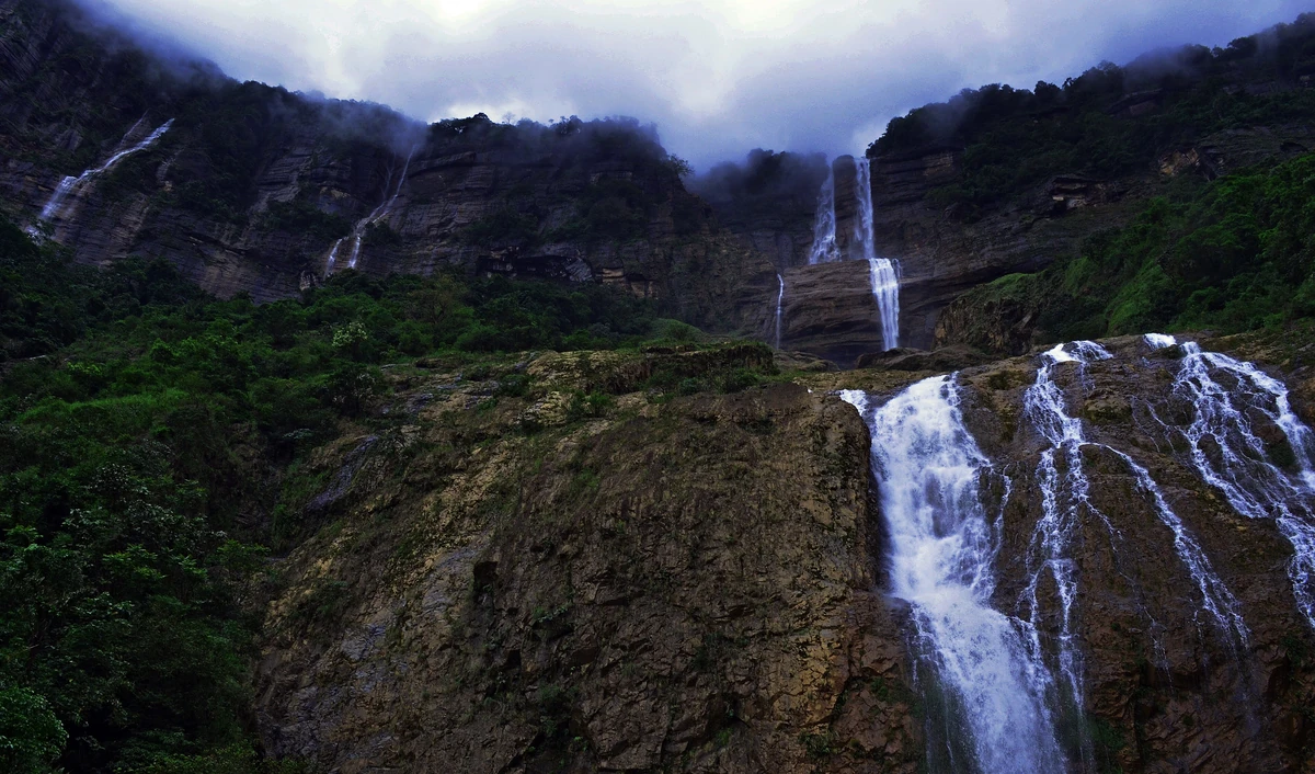 A wide three-tiered waterfall splitting into white streams down a dark rock face below a cloud-capped cliff edge
