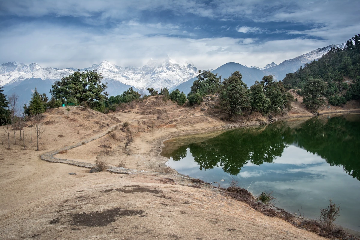 A green forest-ringed lake with a path along its sandy shore and snow-covered mountains rising behind under cloud