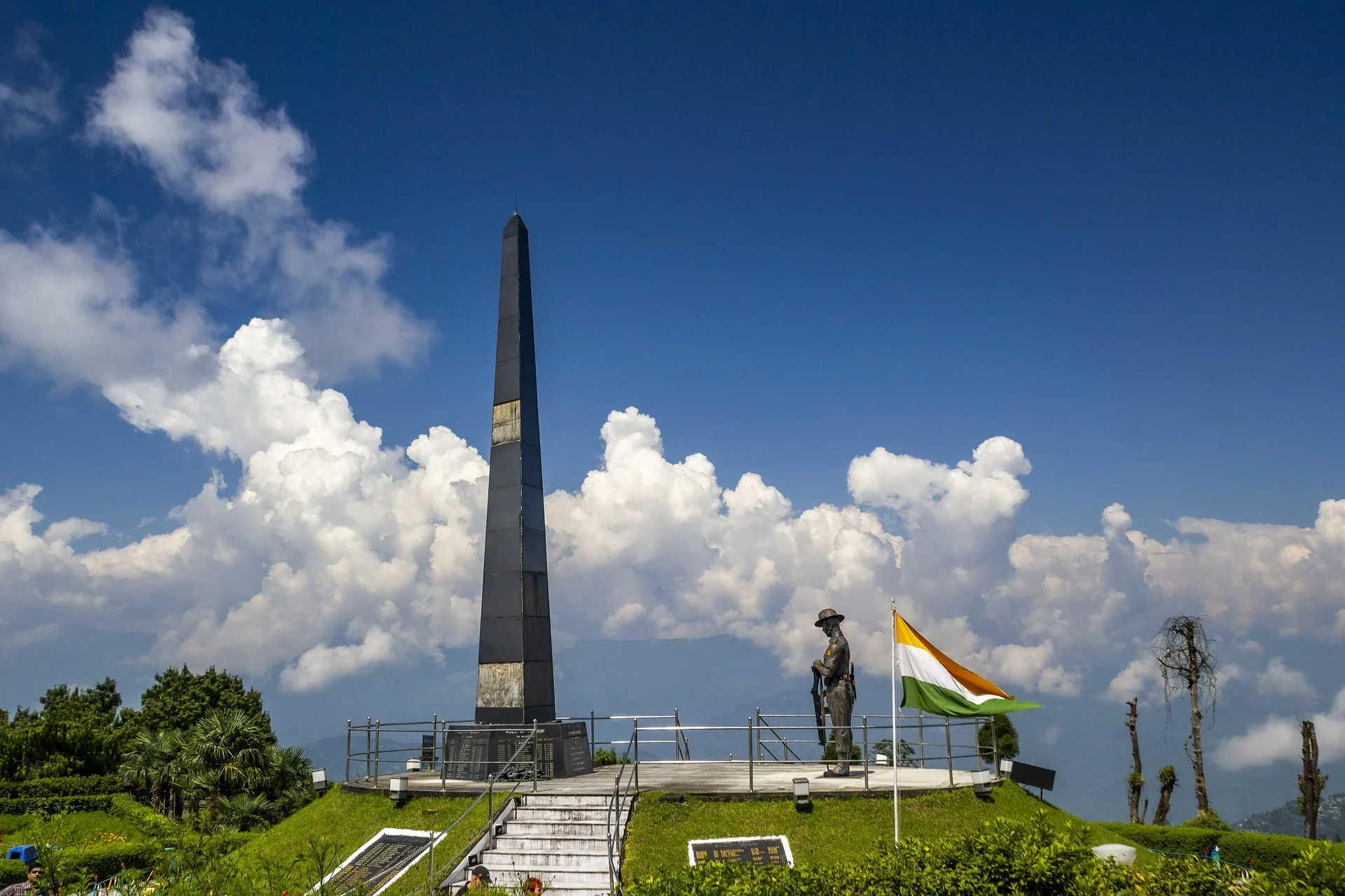 A tall stone obelisk and a soldier's statue on a green garden terrace under a bright sky with towering white clouds