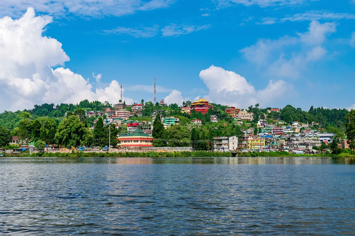 The blue water of Mirik's Sumendu Lake with the town's houses and a monastery on the green ridge beyond under white clouds