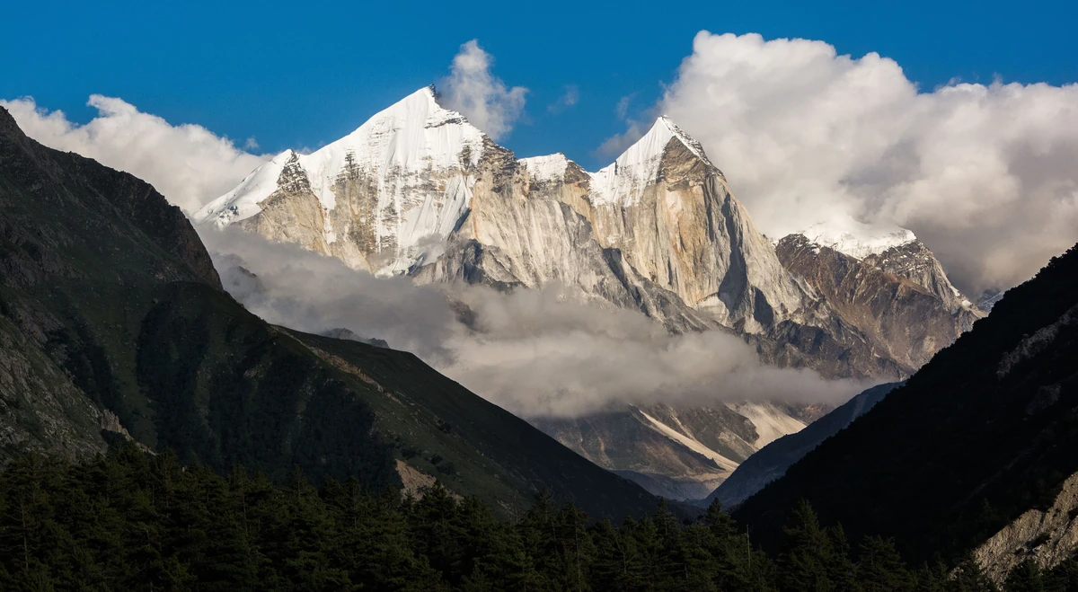 The pale granite summits of the Bhagirathi peaks rising above a green valley with banks of white cloud below