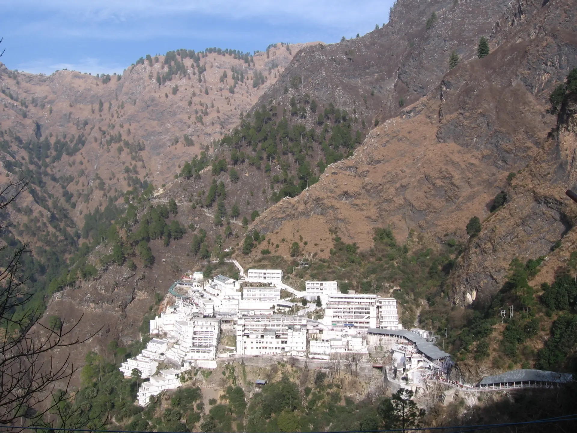 The white buildings of the Vaishno Devi Bhawan on a steep slope of the Trikuta hills, with brown ridges and a blue sky behind