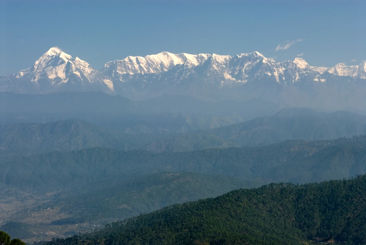 A long line of snow-capped Himalayan peaks rising above layered blue ridges and green hills seen from Kausani