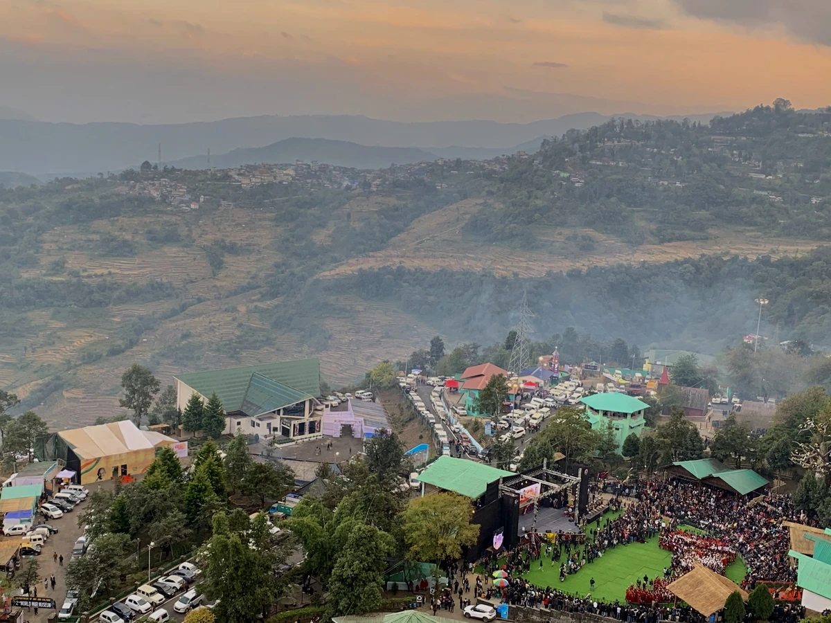 The main arena of the Kisama Heritage Village crowded with spectators during the Hornbill Festival at dusk, with hills behind