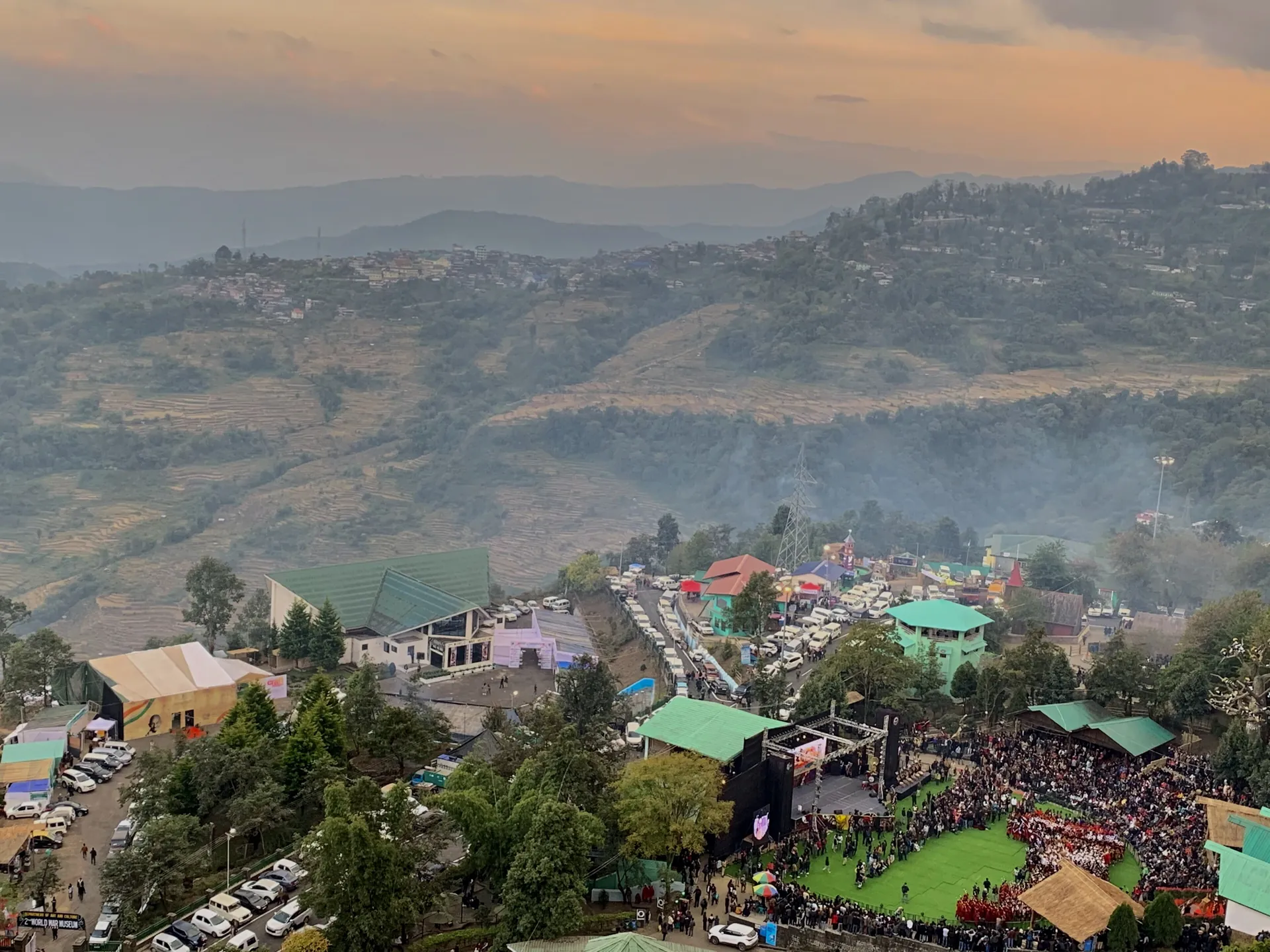 The main arena of the Kisama Heritage Village crowded with spectators during the Hornbill Festival at dusk, with hills behind