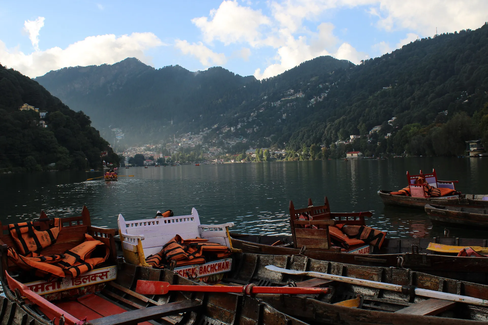 Rows of boats with red cushioned seats moored on Nainital Lake, with forested hills across the water under a blue sky