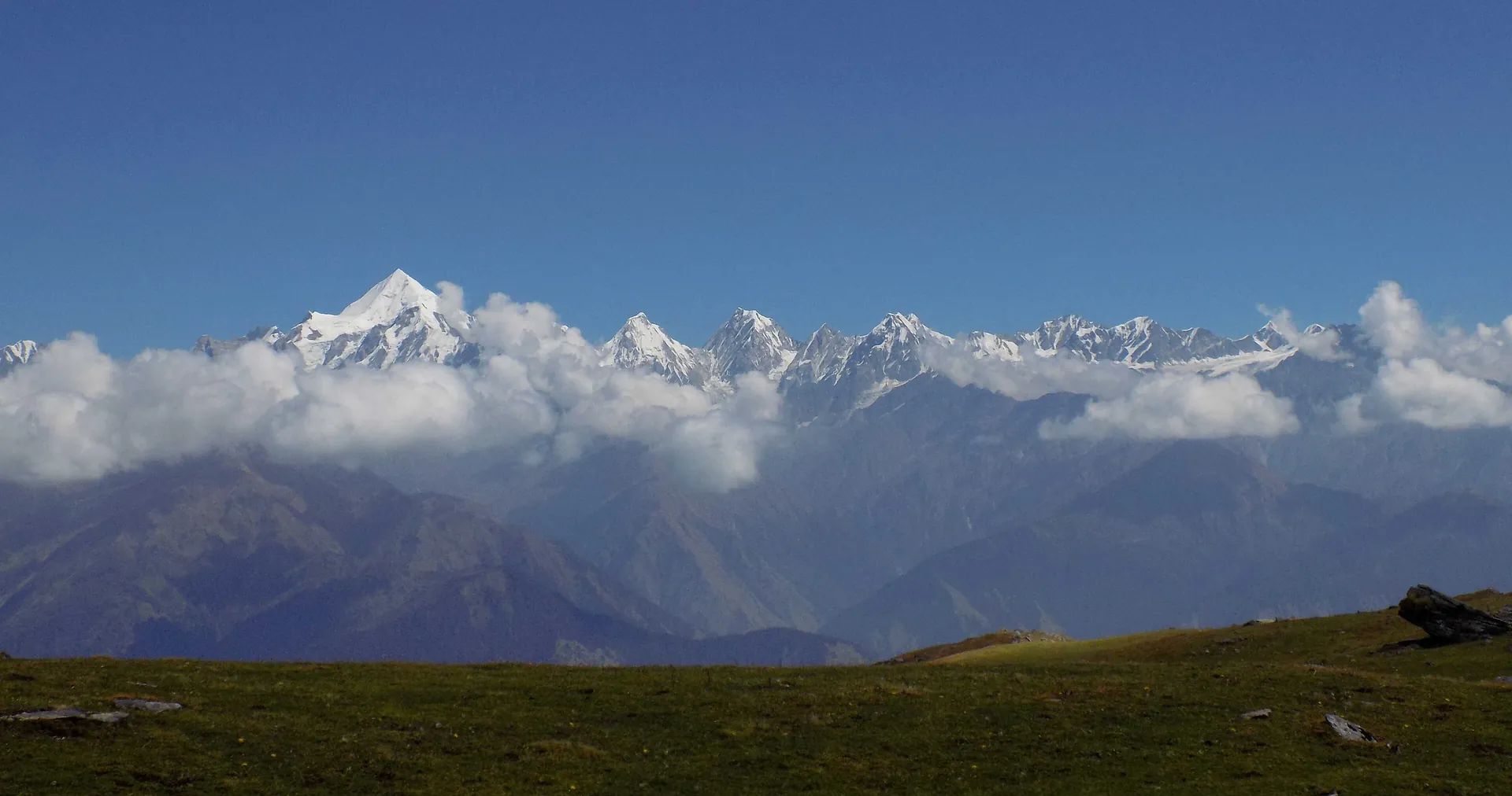 The snow-covered Panchachuli peaks along the horizon above banks of cloud and dark foothills seen from a meadow near Munsiyari