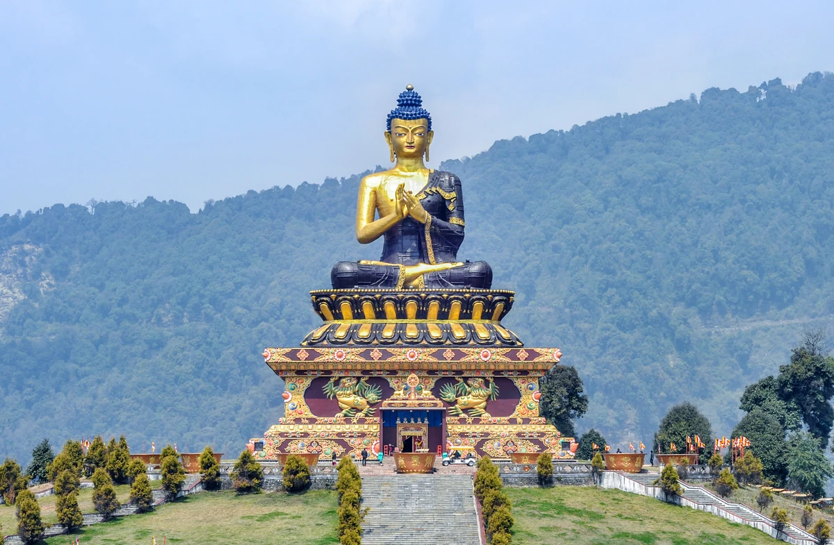 The seated golden and dark-blue Buddha statue on a stepped, richly painted base against a forested hillside and a cloudy sky