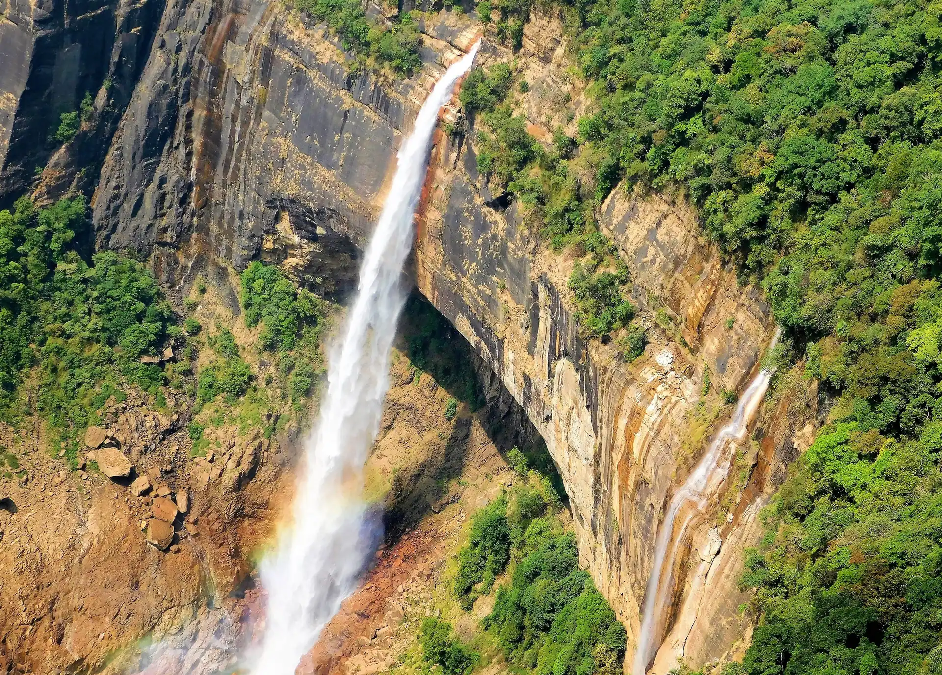 A tall narrow waterfall plunging down a sheer cliff face beside green forest, with a faint rainbow at its base