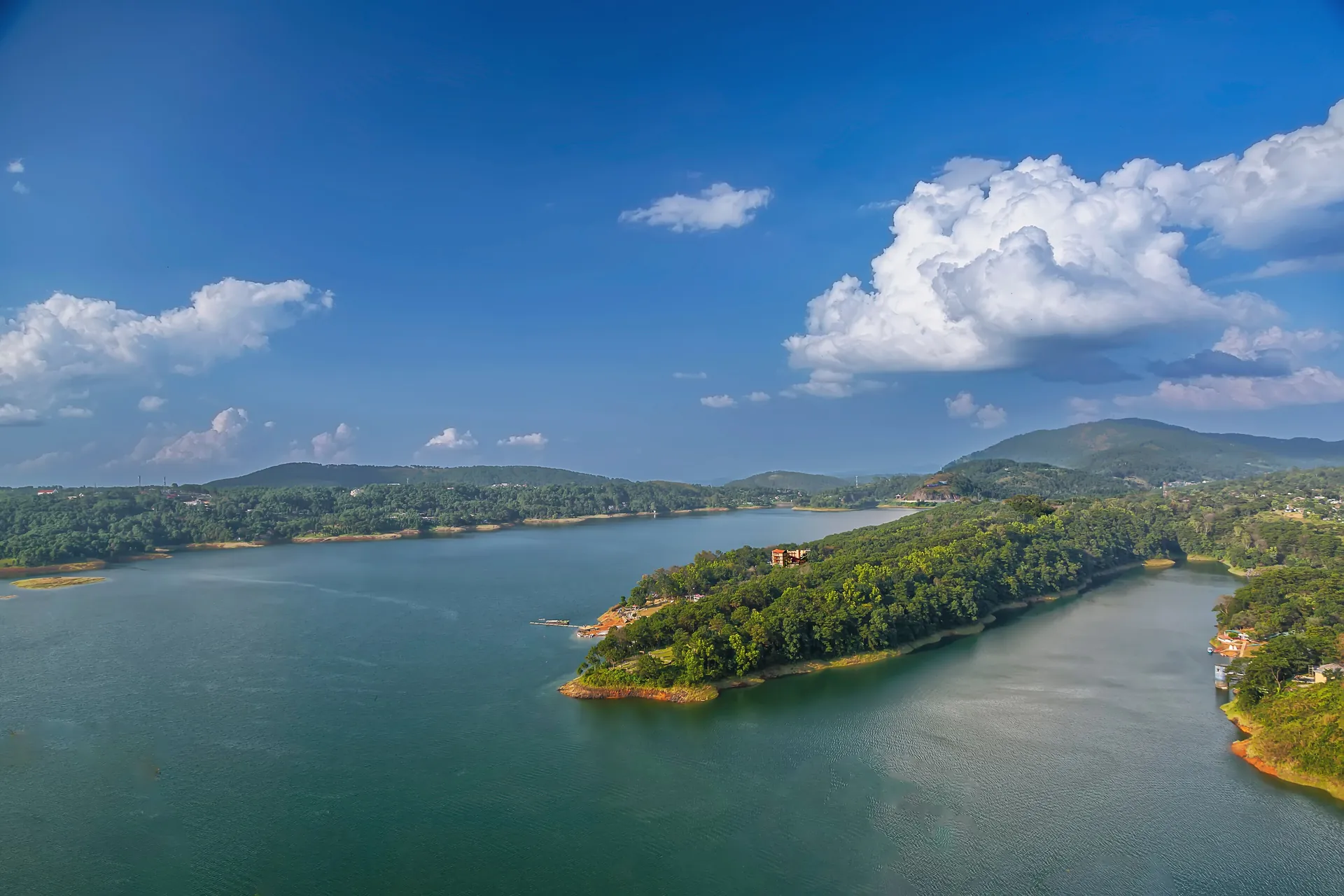 An aerial view of Umiam Lake with its forested peninsulas and green water under a blue sky with white cloud