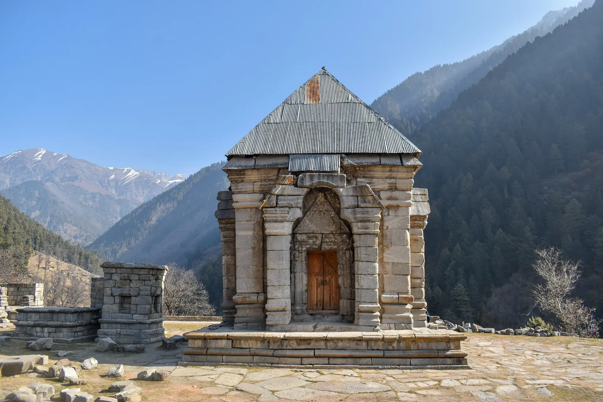 A small stone temple with a grey pyramid roof and a carved doorway standing on a stone platform below forested mountains at Naranag