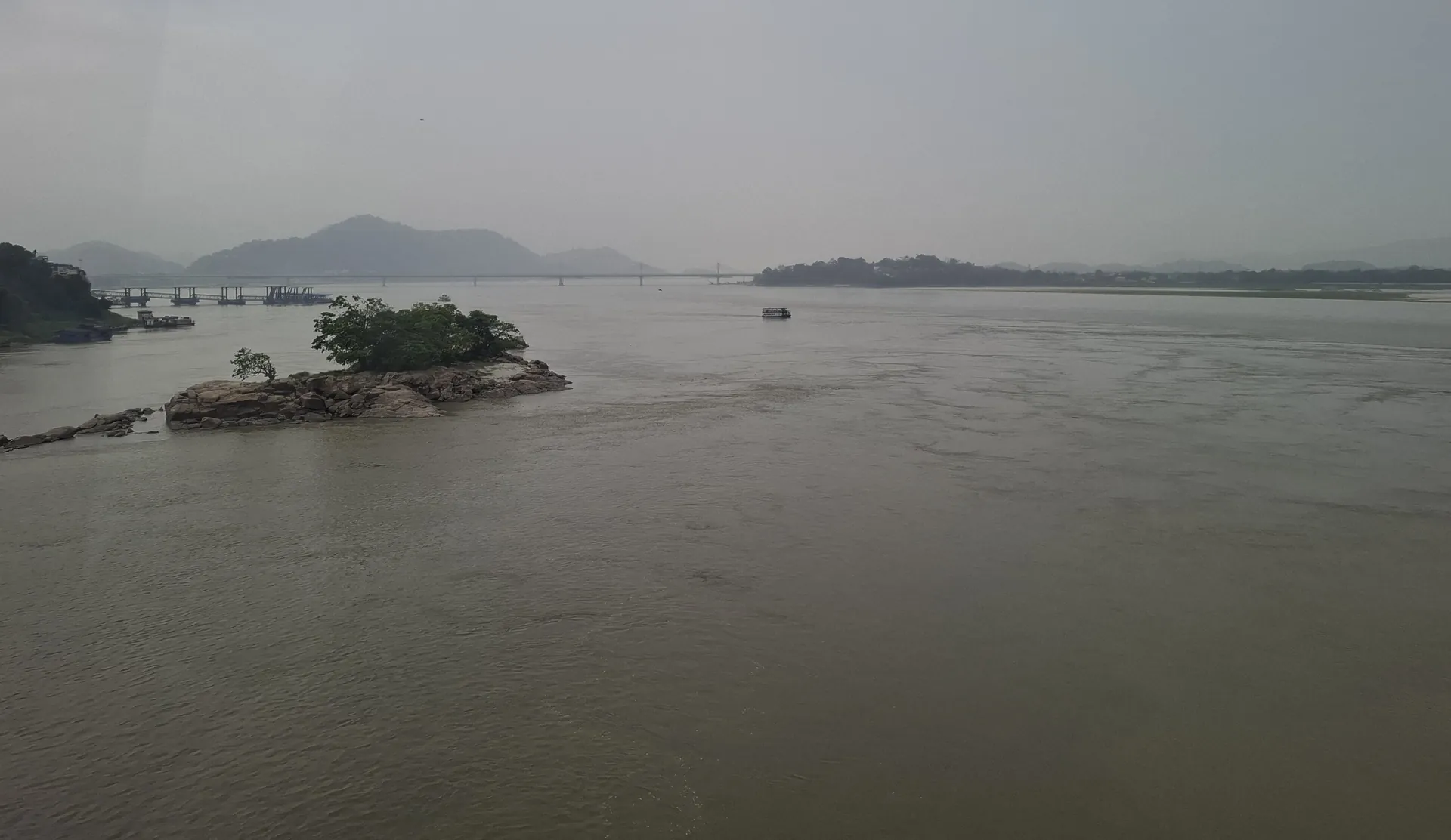 A wide grey stretch of the Brahmaputra with a small wooded rocky island and low hills in the haze beyond