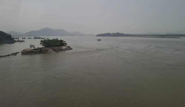 A wide grey stretch of the Brahmaputra with a small wooded rocky island and low hills in the haze beyond