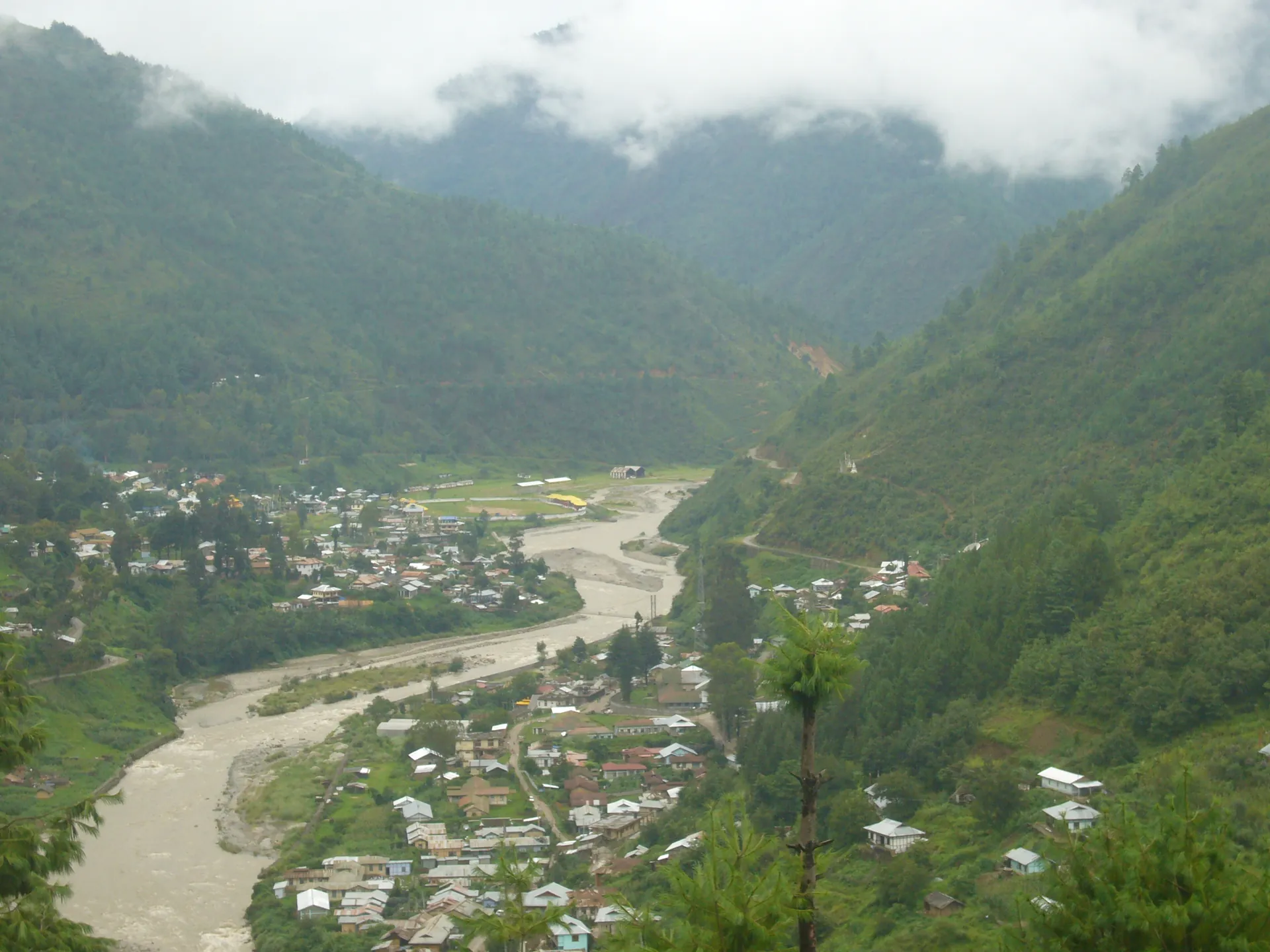 A pale river winding through the green valley floor at Dirang, with houses on the slopes and cloud over the hills