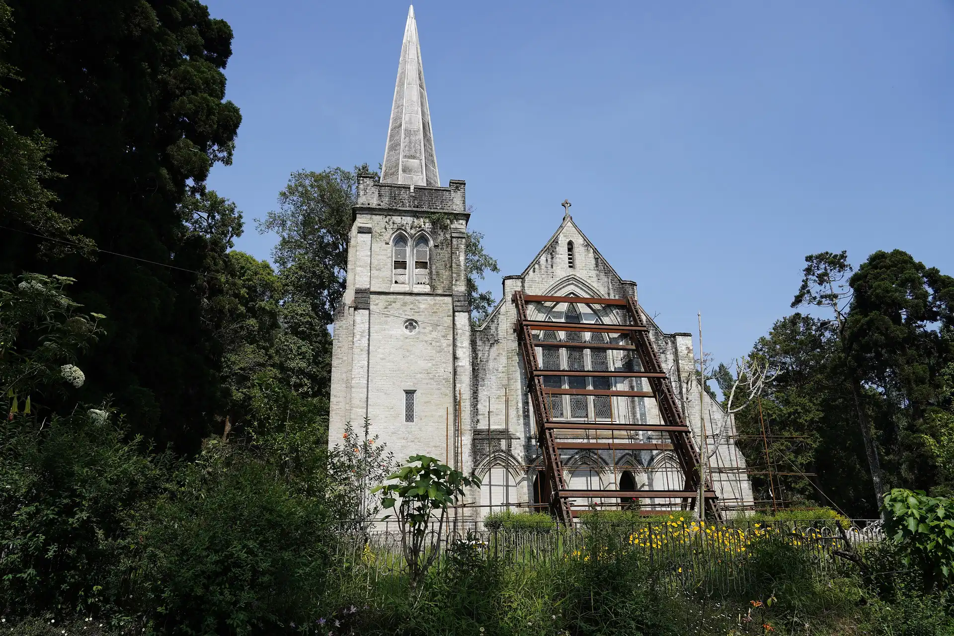 A grey stone chapel with a tall spire and a pointed gable among tall trees under a blue sky
