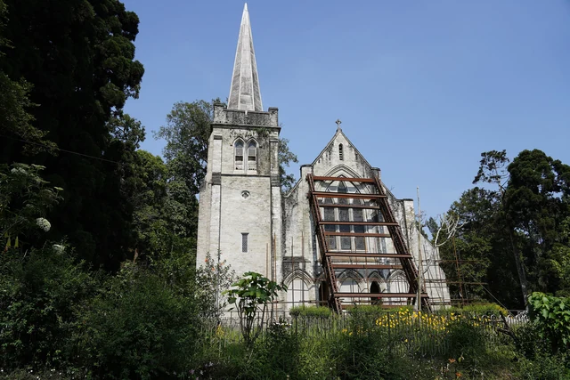A grey stone chapel with a tall spire and a pointed gable among tall trees under a blue sky