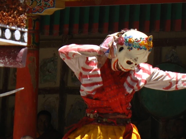 A masked dancer in a white mask with a flower crown, a red and white striped robe and yellow skirt at a monastery festival