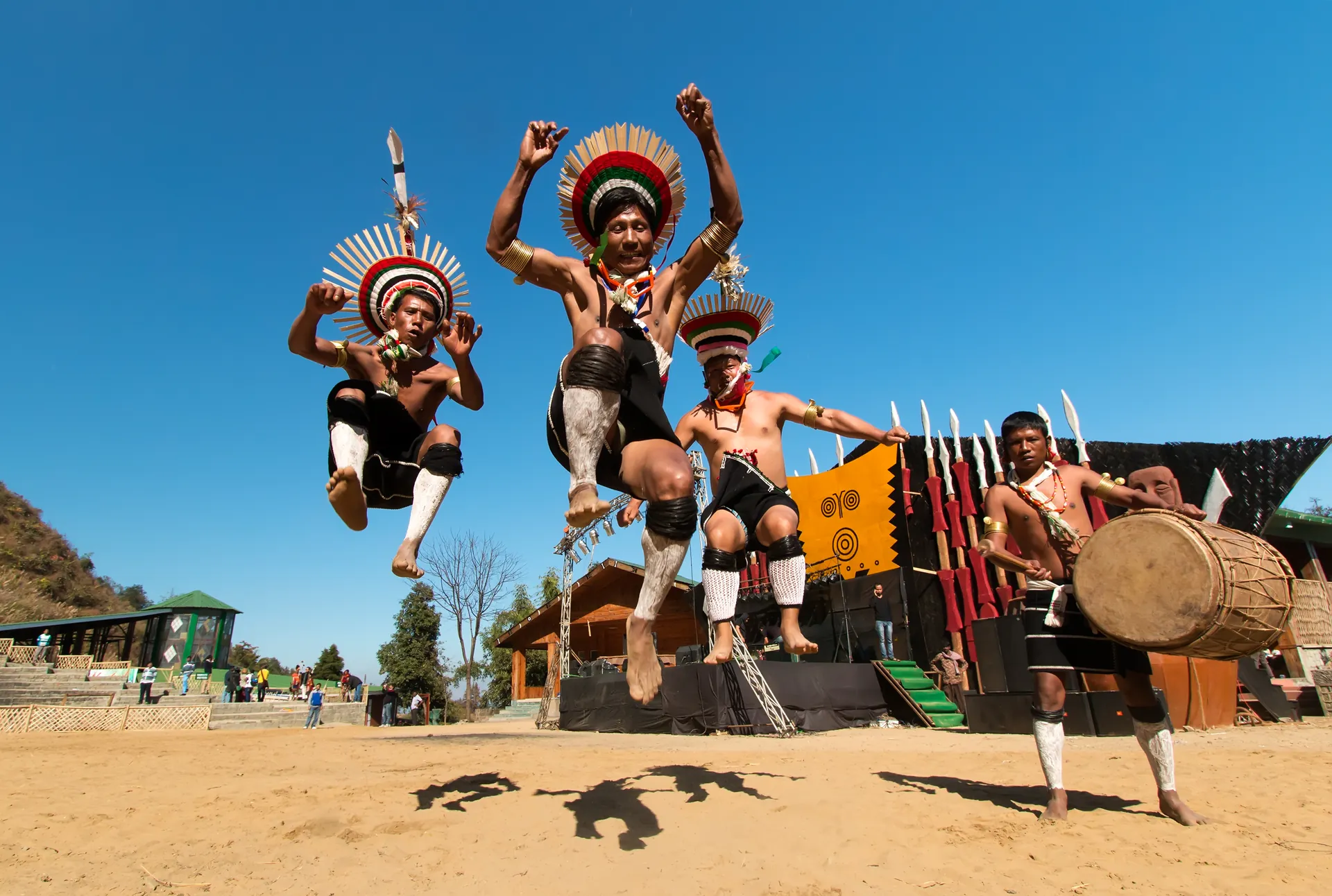 Naga dancers in feathered headdresses leaping into the air beside a log drum on a dusty festival ground under a blue sky