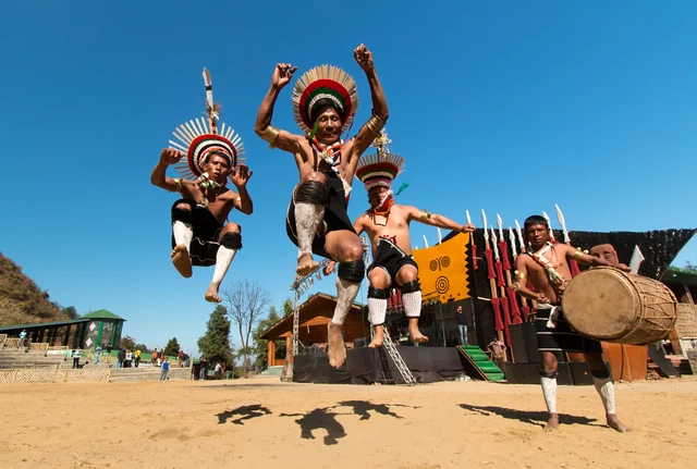 Naga dancers in feathered headdresses leaping into the air beside a log drum on a dusty festival ground under a blue sky