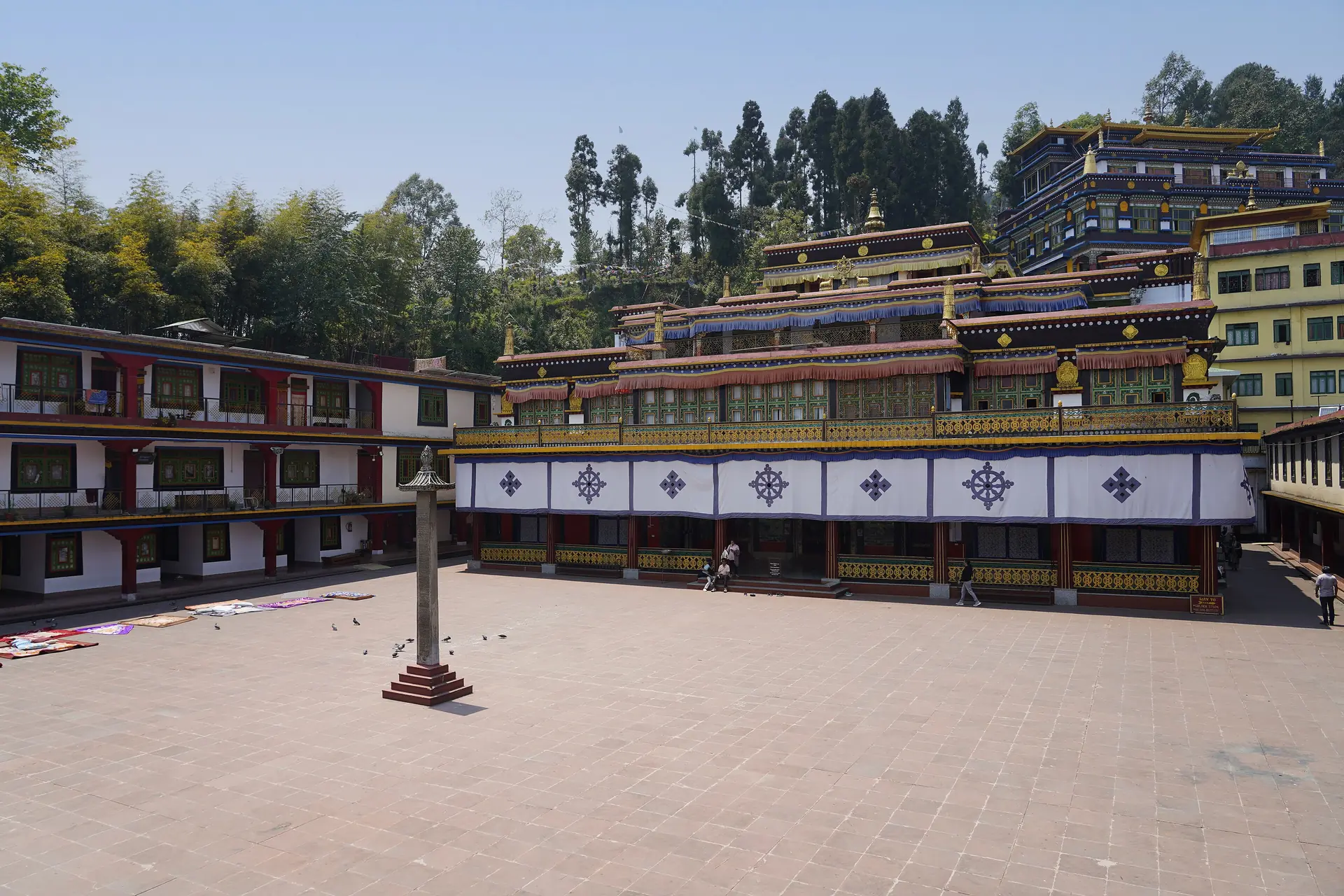 The wide brick courtyard of Rumtek Monastery with its colonnaded white and red monastery building behind, among tall trees