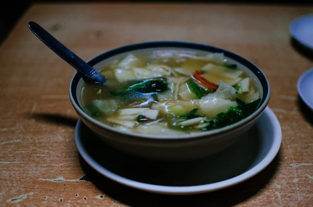 A bowl of thukpa, a noodle soup with flat noodles, green leaves and slices of carrot, with a spoon, on a wooden table