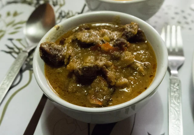 A white bowl of dark pork stew cooked with fermented soybeans on a table beside a spoon and fork
