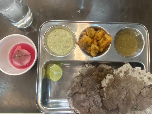 A steel tray holding a dark round flatbread, fried potato pieces, two chutneys, a slice of lime and a cup of pink drink