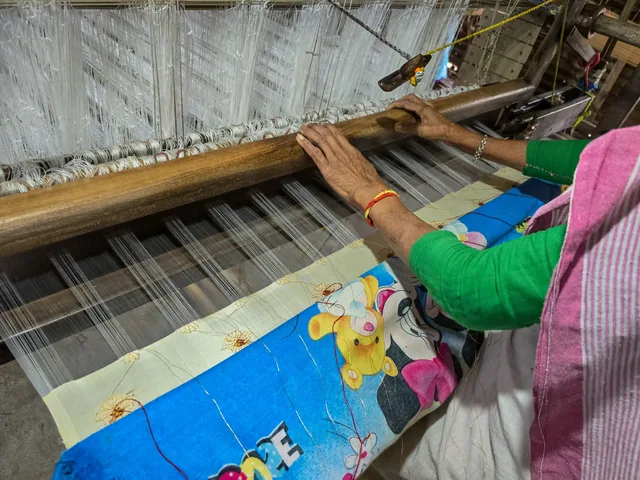 A weaver's hands working threads on a handloom, with a half-woven blue and pink cloth in front