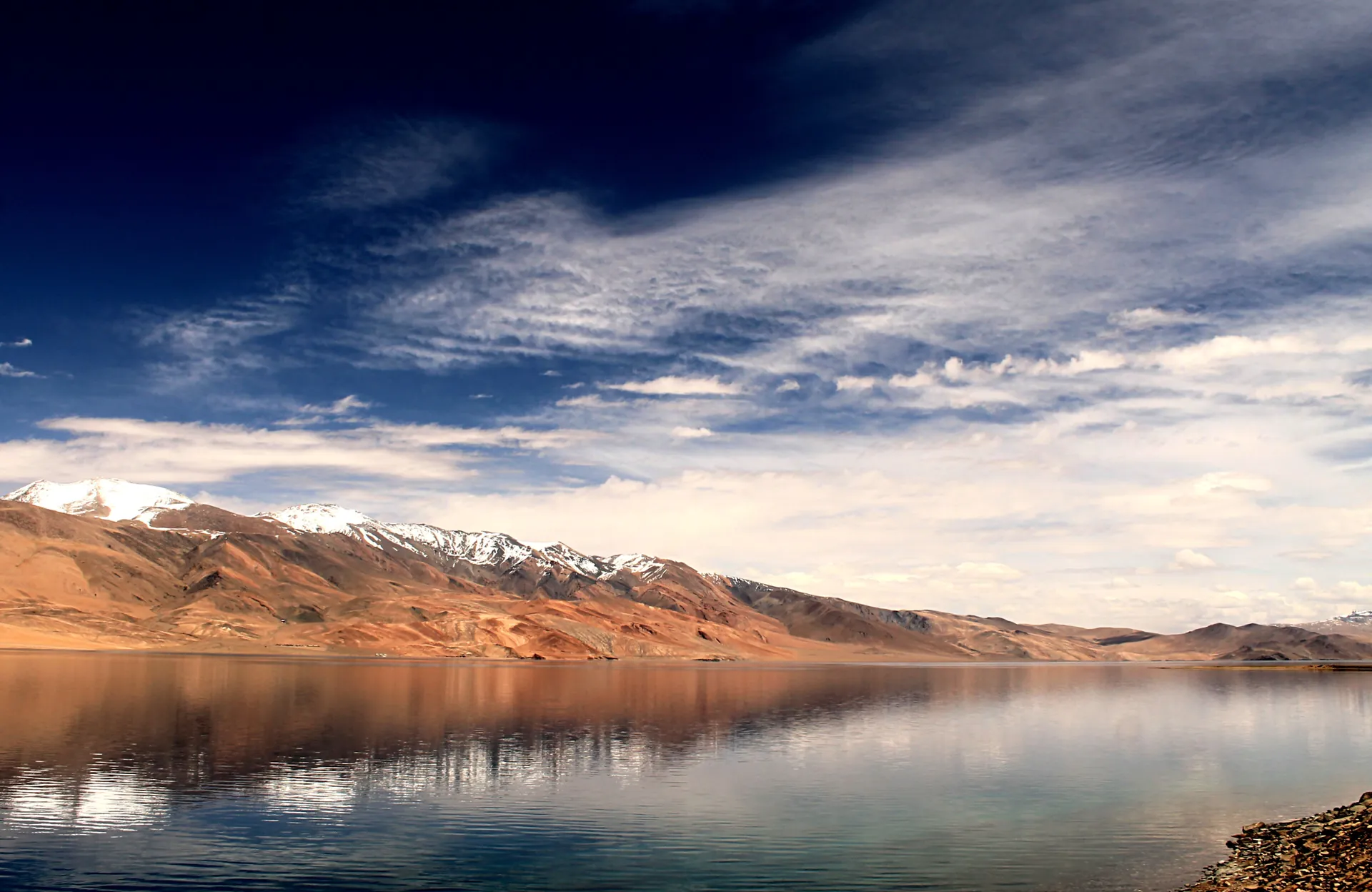 Tso Moriri's still blue water reflecting brown hills and snow peaks under a dark sky streaked with cloud