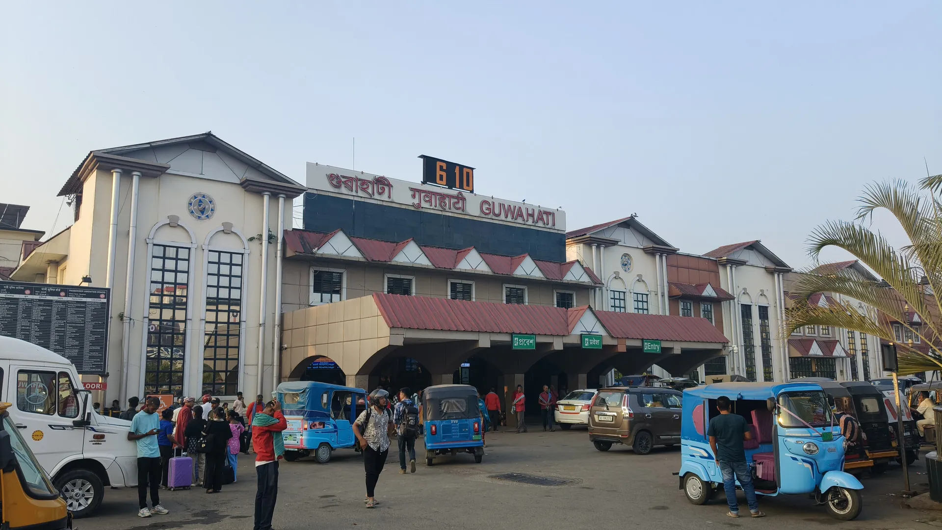 The front of Guwahati railway station at Paltan Bazaar, with blue auto-rickshaws and people outside its red-roofed entrance