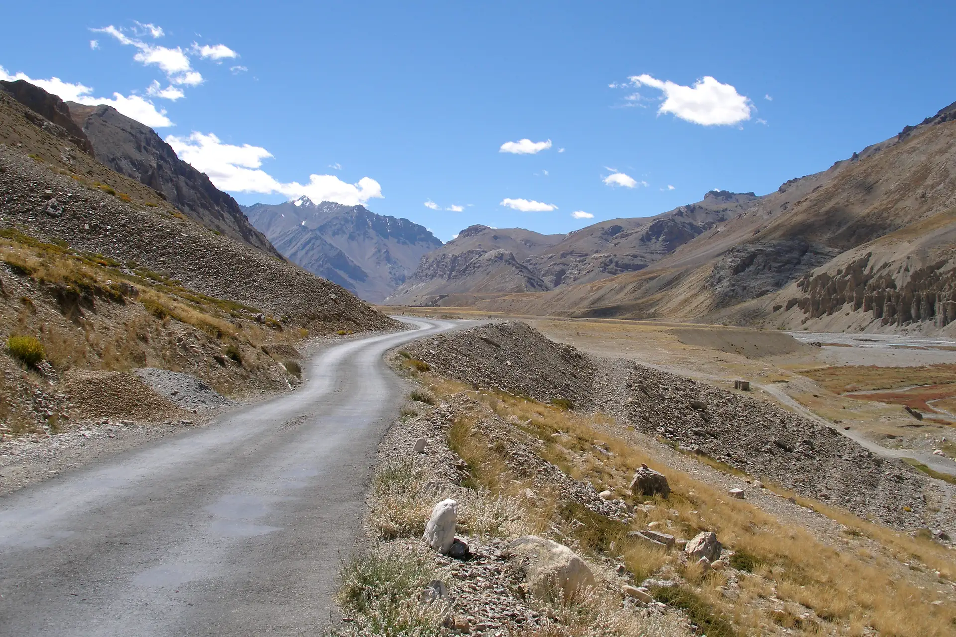 A narrow paved road winding along a barren valley of brown and grey mountains under a blue sky with scattered clouds