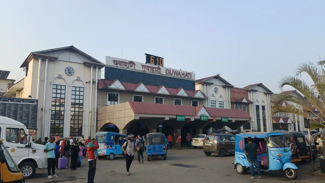 The front of Guwahati railway station at Paltan Bazar, with blue auto-rickshaws and people outside its red-roofed entrance
