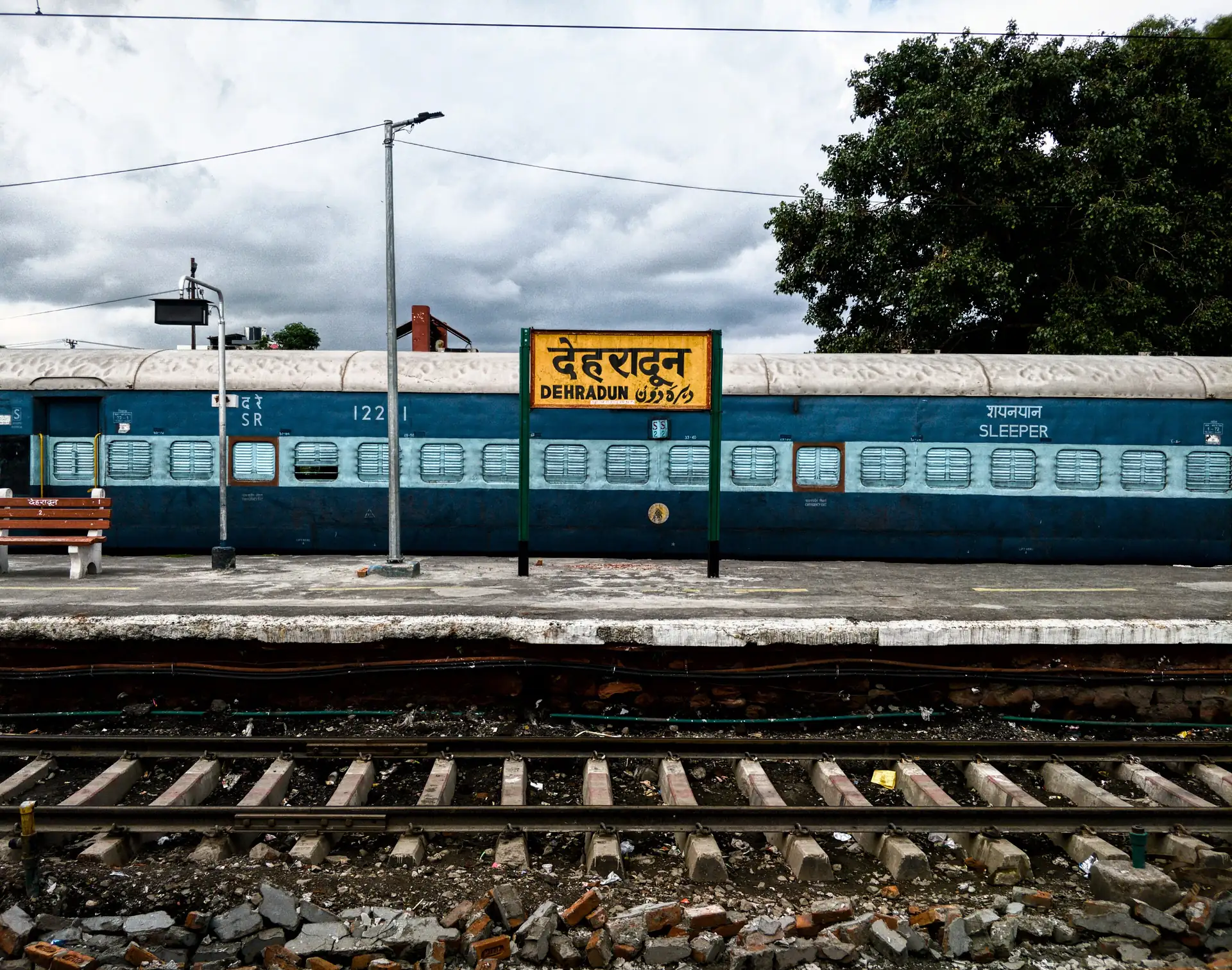A blue sleeper-class train standing at a platform beside a yellow Hindi and English station board reading Dehradun