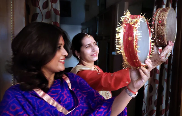Two women in festive clothes holding up a decorated sieve and a puja plate towards the night sky