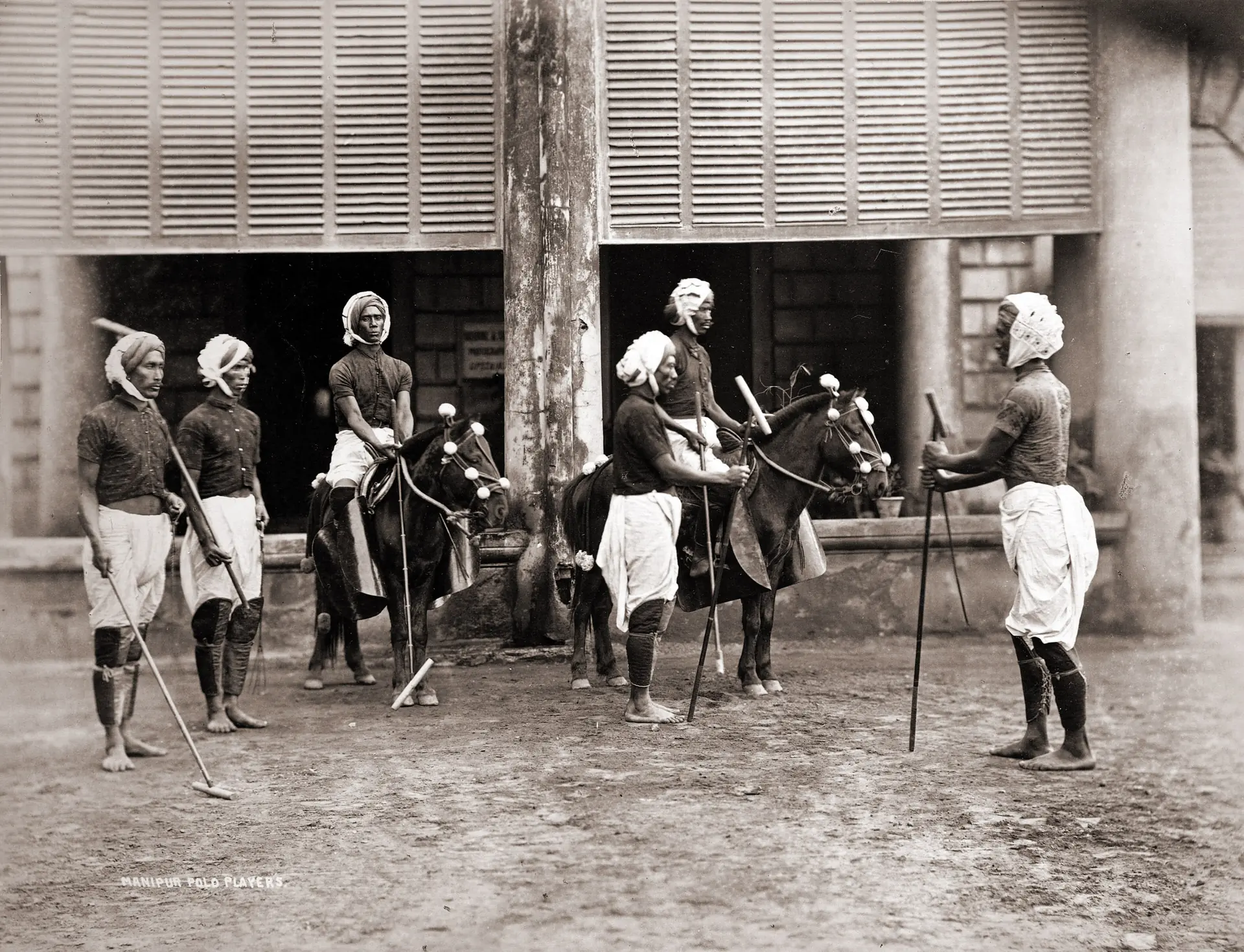 Manipuri polo players with their sticks and ponies outside a timber building, photographed in 1875