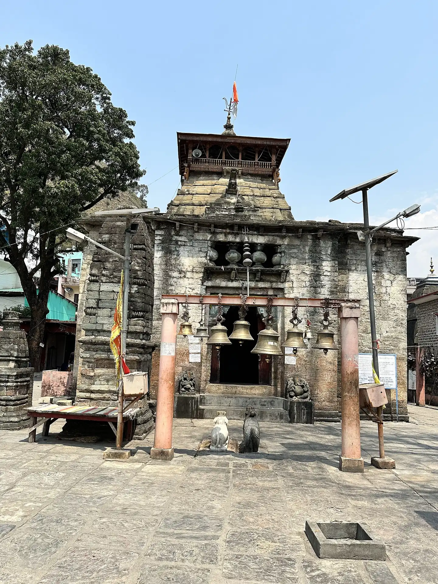 The stone entrance gate of the Bagnath temple with hanging bells and a small tower behind it on a paved forecourt