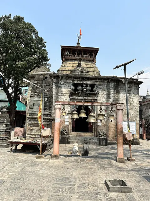 The stone entrance gate of the Bagnath temple with hanging bells and a small tower behind it on a paved forecourt
