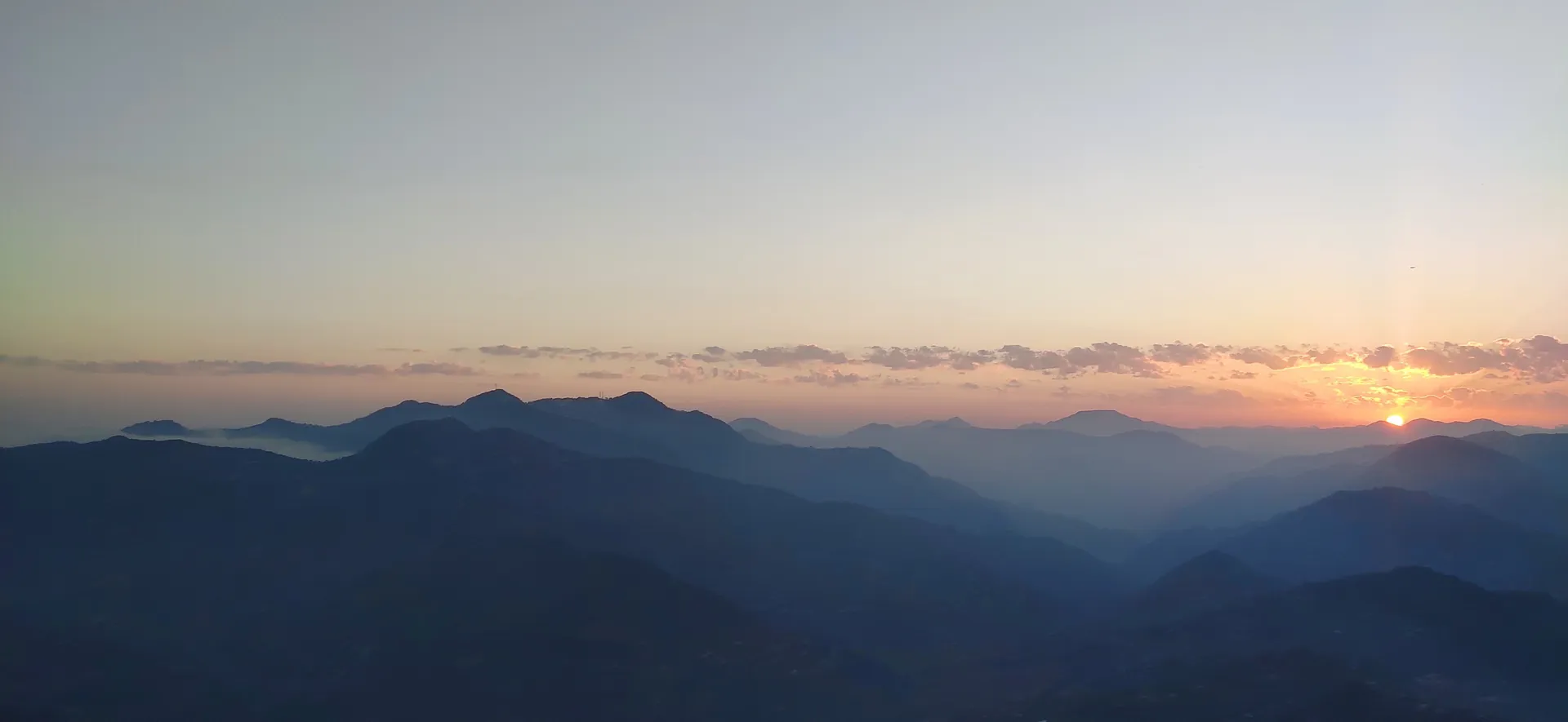 A wide orange glow along the horizon above layered dark ridges at dusk from Mukteshwar