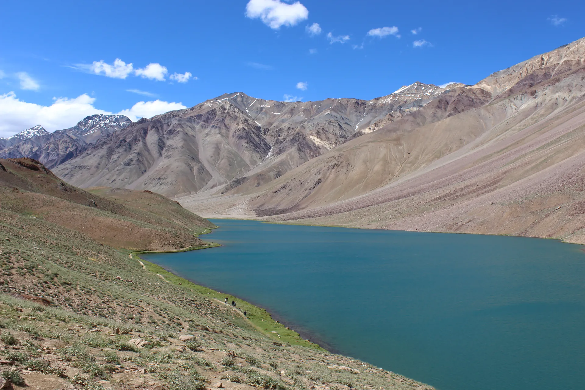Chandratal, a curved blue lake below bare brown and grey scree slopes in Lahaul, under a blue sky