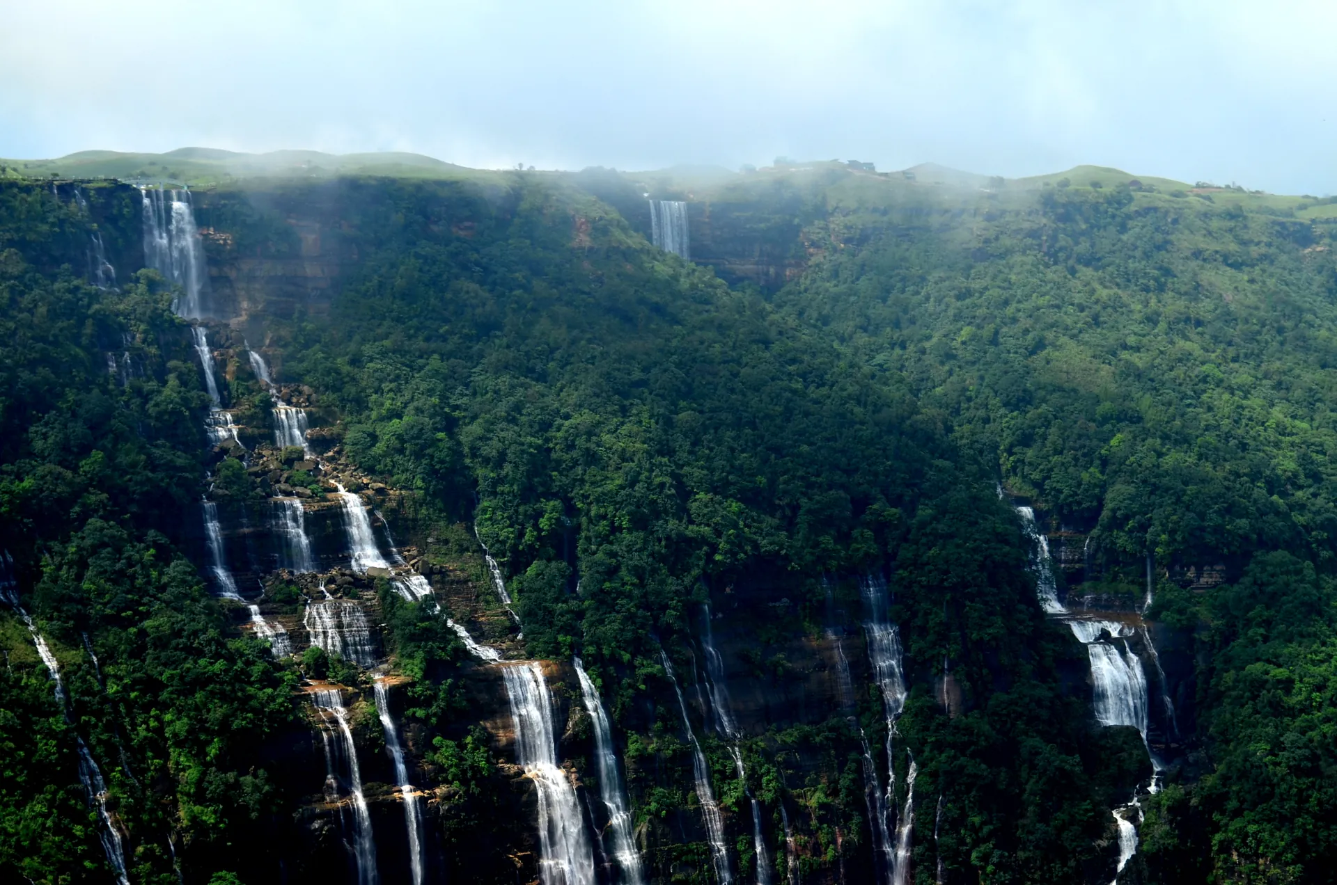 A line of thin white waterfalls dropping down a long green cliff face in several separate streams under a hazy sky