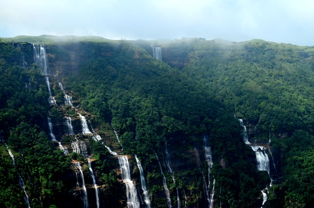 A line of thin white waterfalls dropping down a long green cliff face in several separate streams under a hazy sky