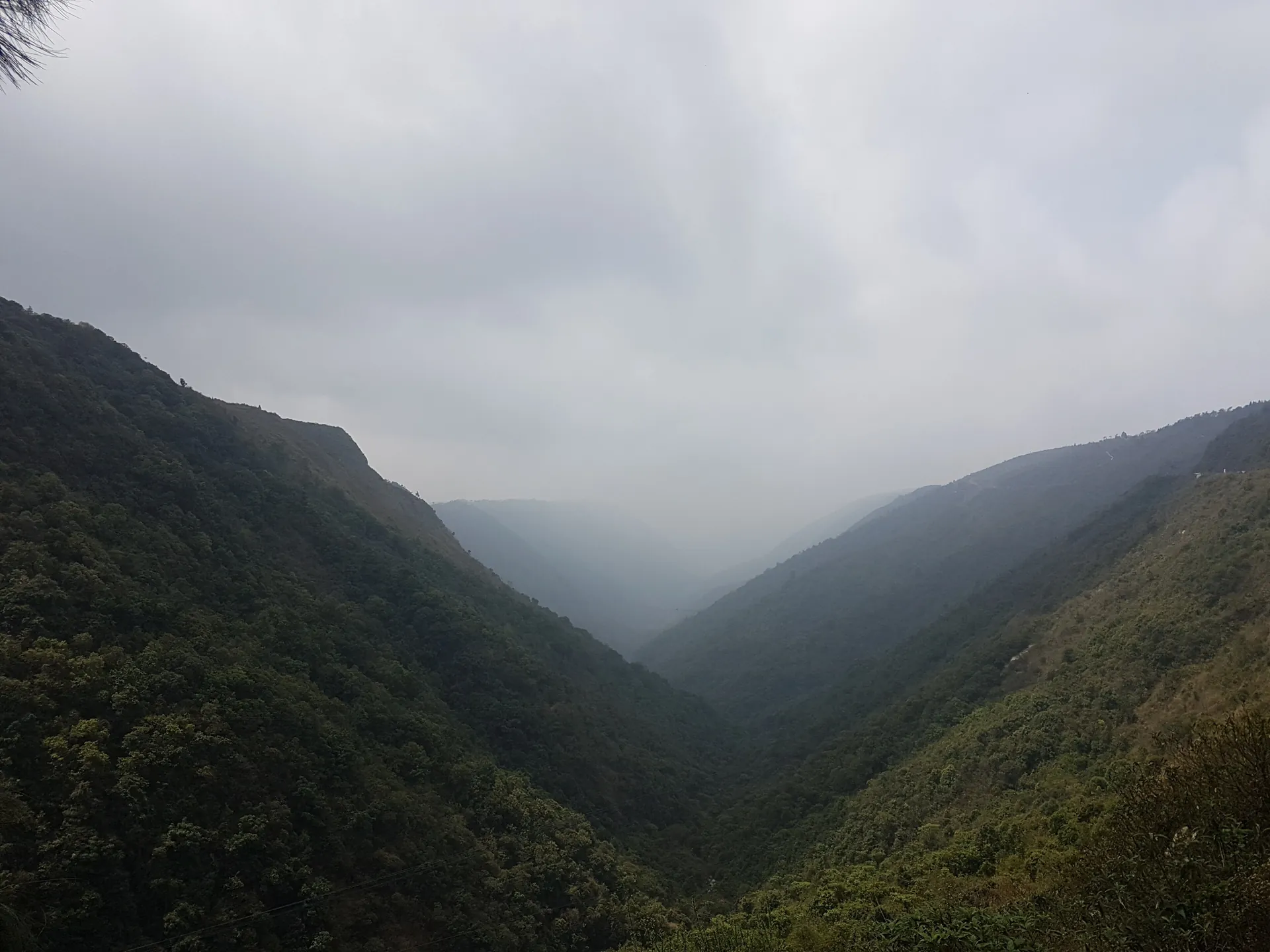A deep, steep-sided green valley filled with low grey cloud seen from the Mawkdok Dympep viewpoint near Cherrapunji