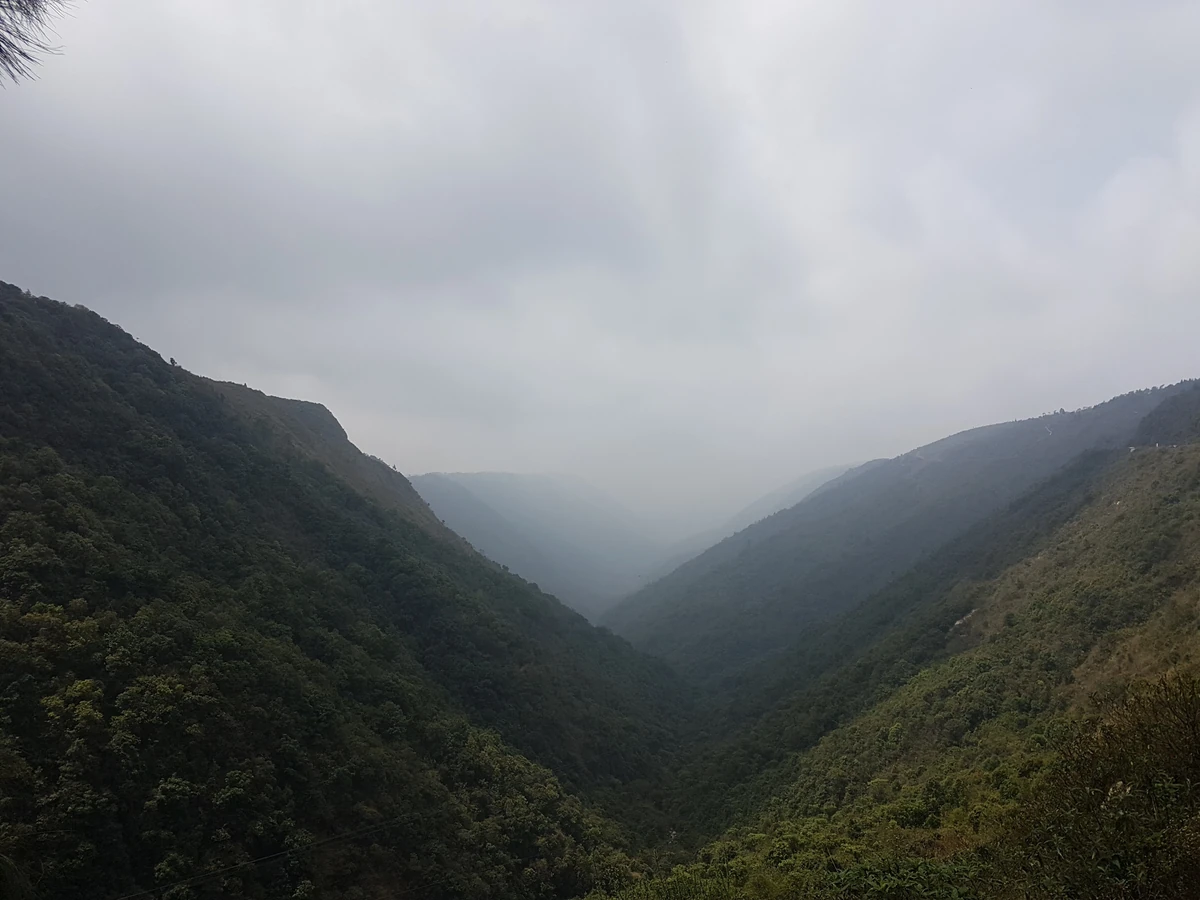 A deep, steep-sided green valley filled with low grey cloud seen from the Mawkdok Dympep viewpoint near Cherrapunji