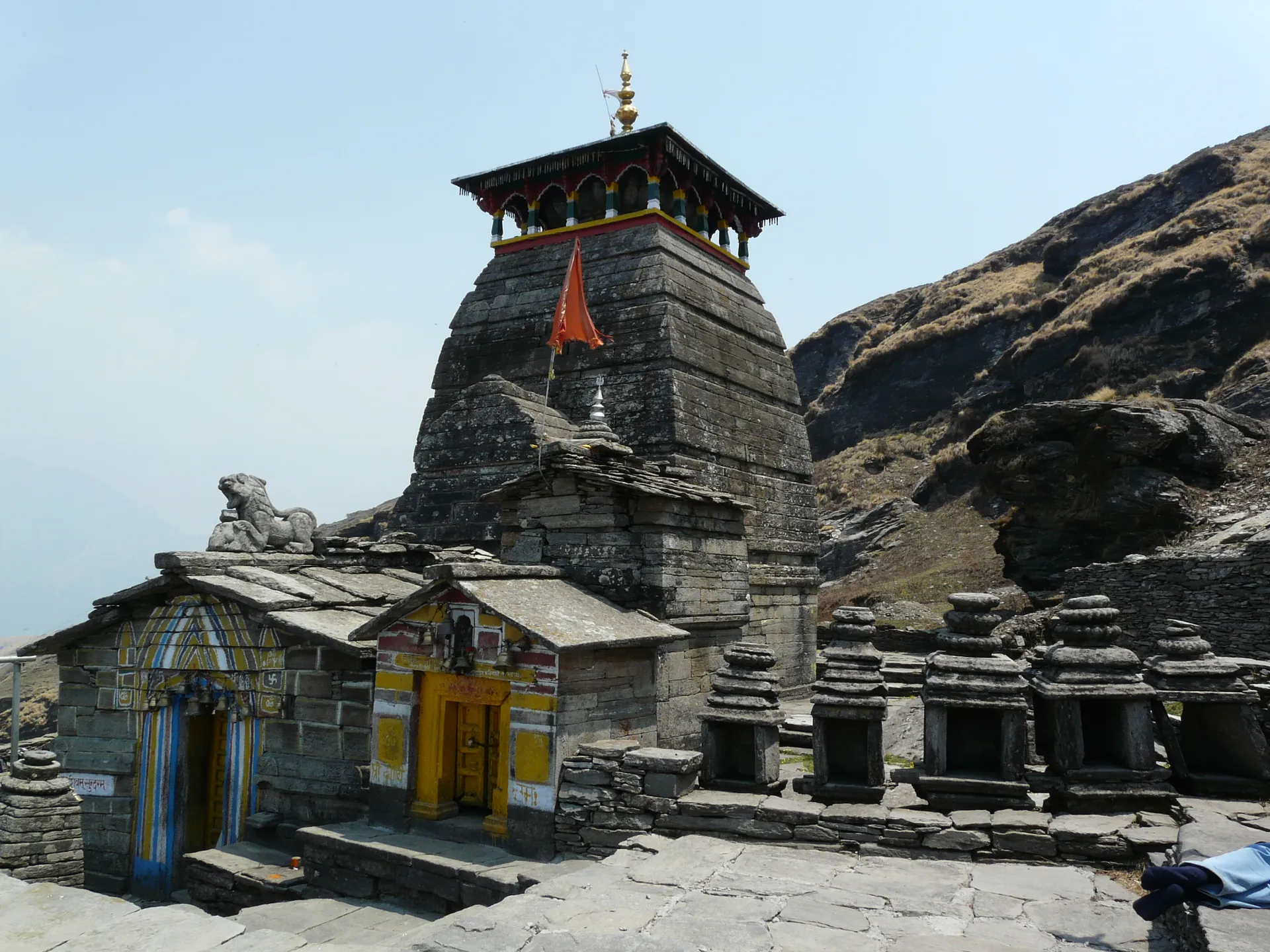 The grey stone Tungnath Temple with a small shrine in front of it, on a rocky ridge under a clear blue sky