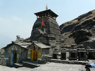 The grey stone Tungnath Temple with a small shrine in front of it, on a rocky ridge under a clear blue sky