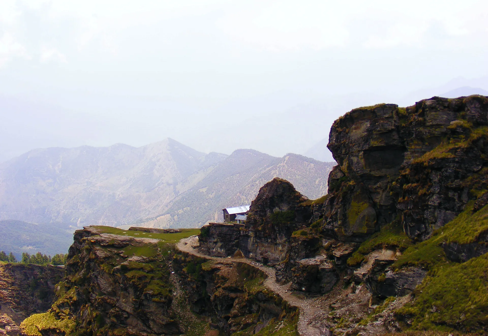 A path winding along a rocky ridge on the Chandrashila trek with layered mountain ranges in a haze behind