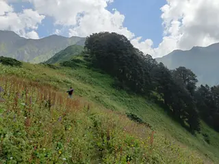 A trekker walking up a green slope towards Dehnasar Lake under a cloudy sky