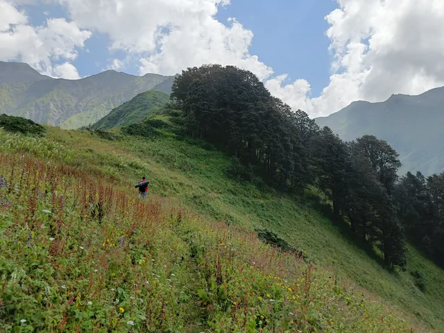 A trekker walking up a green slope towards Dehnasar Lake under a cloudy sky