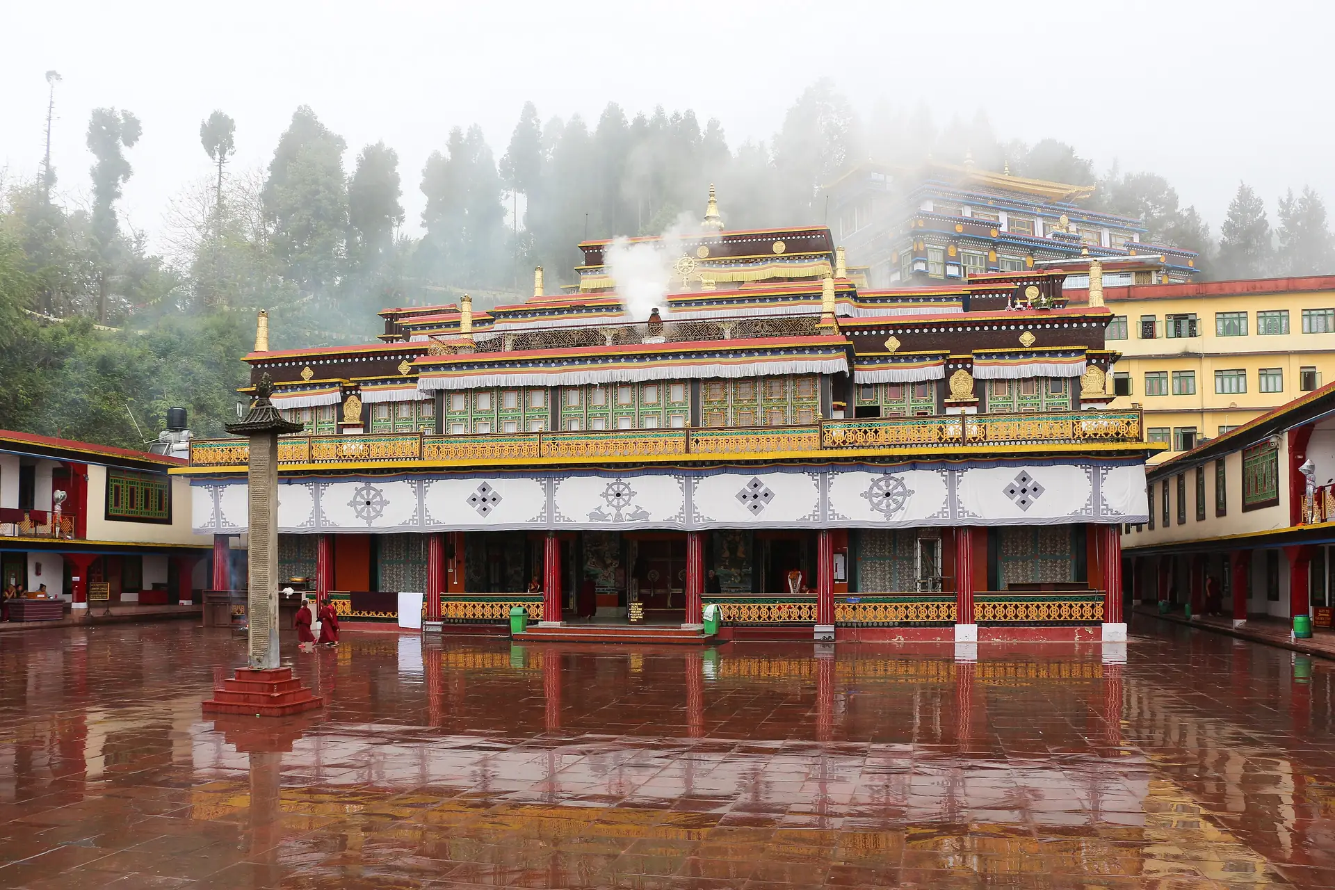 The red-pillared prayer hall of Rumtek Monastery across a wet courtyard, with mist and trees behind it